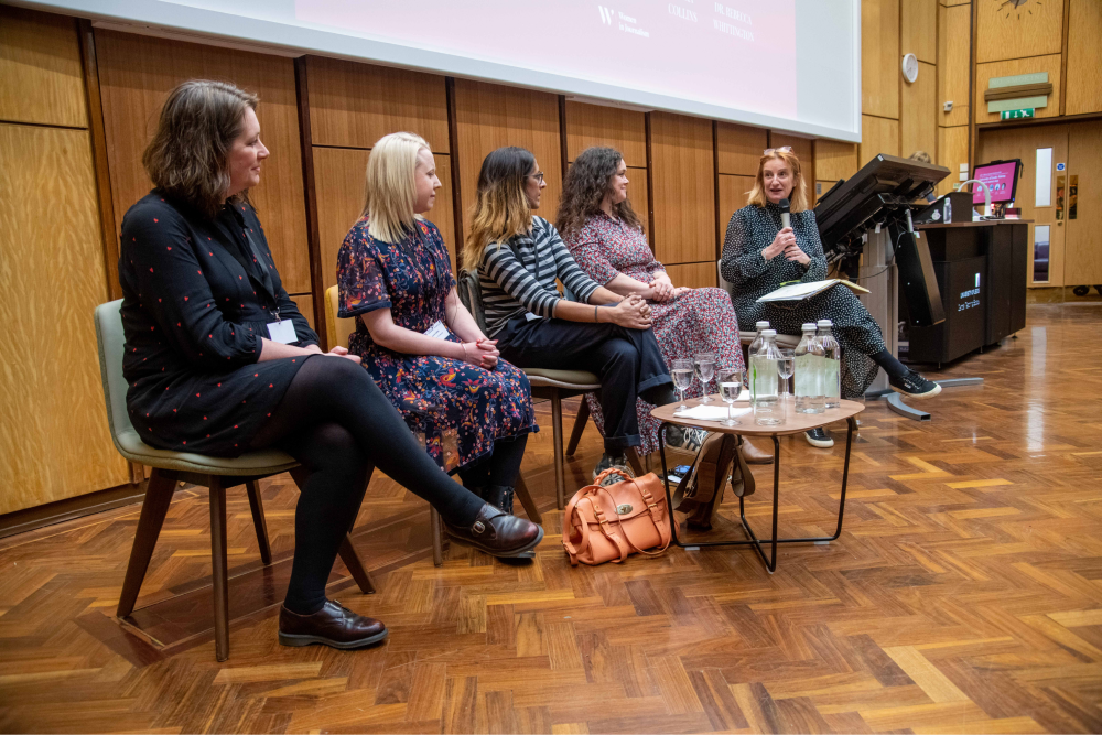 Alison sits alongside fellow journalists in a line, holding a microphone as she asks a question
