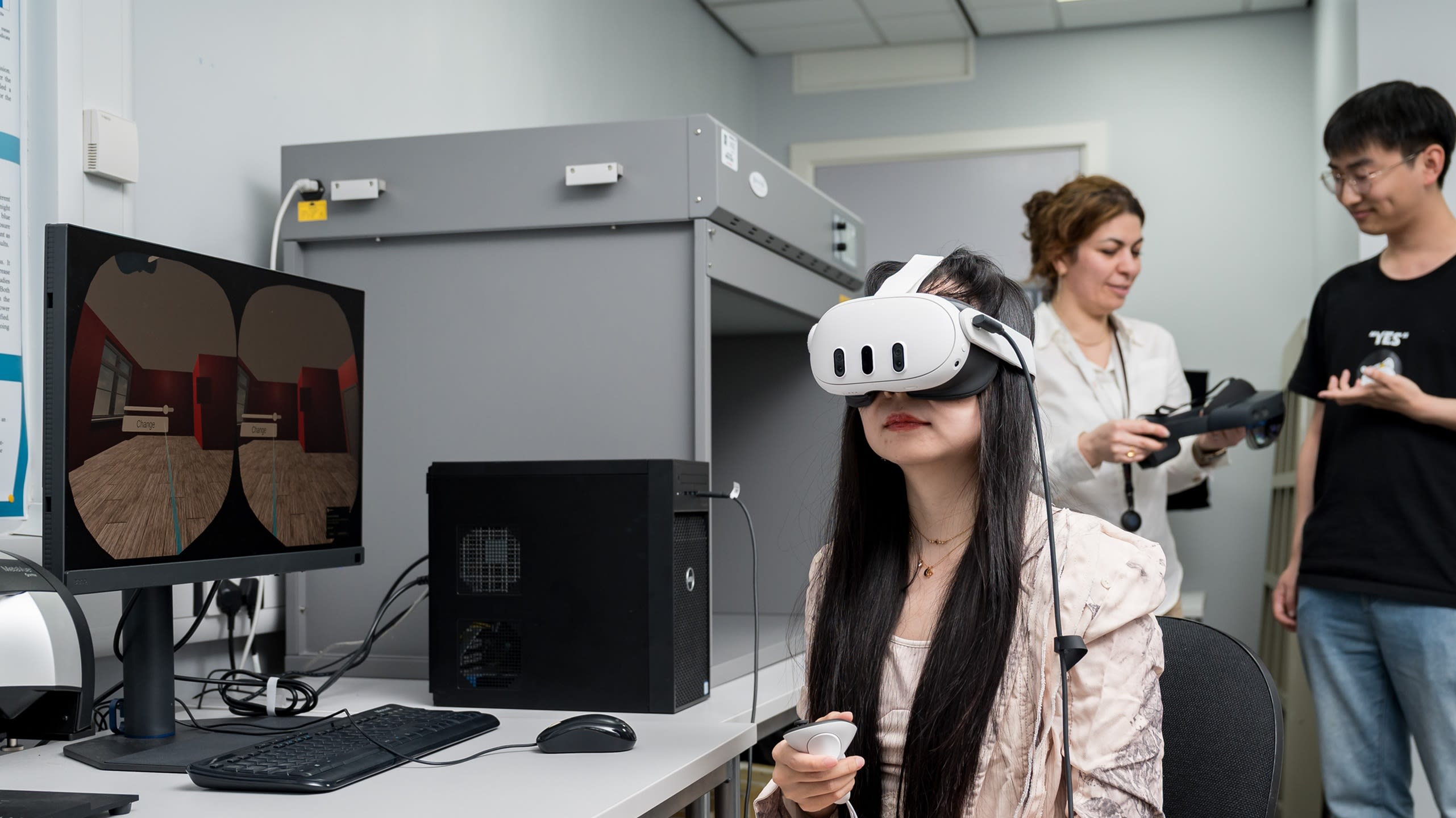 A person wearing a VR headset in a lab, sat at a desk with computer. Two researchers are talking in the background.
