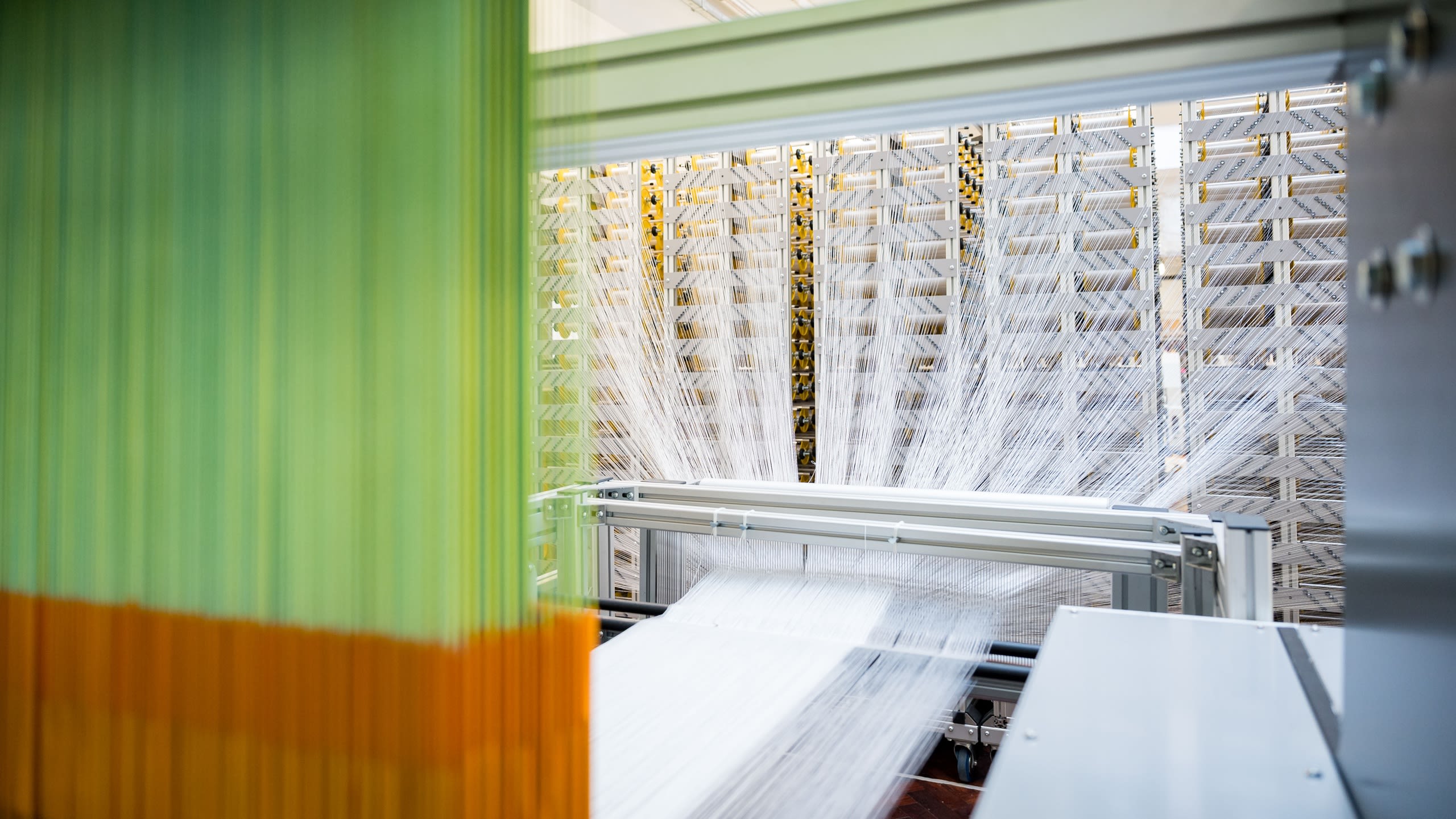 A large weaving machine weaving white threads, with green and orange threads hanging down in the foreground.