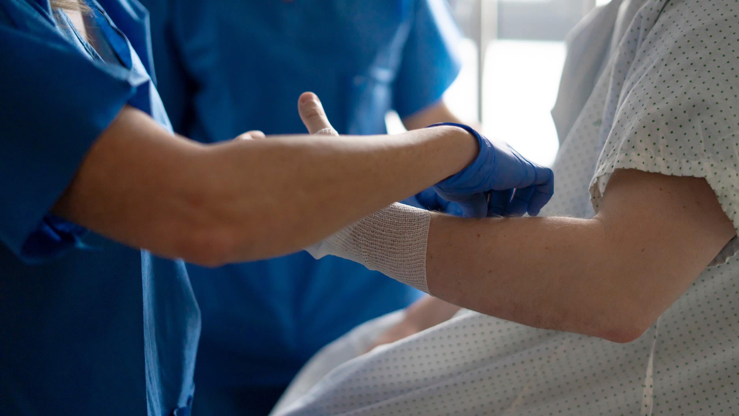 A patient in a gown having their arm bandaged by two medical staff wearing blue gowns.