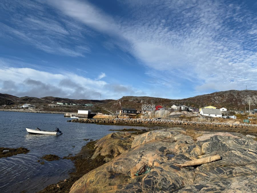View from a rock in the sea of a small village with a handful of houses sat on the rocky coastline. A small boat floats in the water.