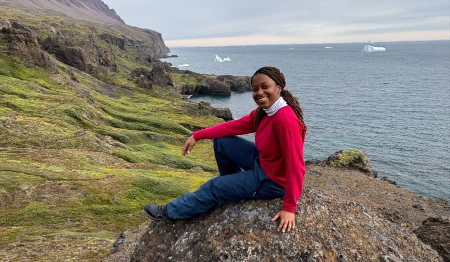 Sanola sits smiling on a large rock on a grassy area that leads to a jagged and rocky coastline. The sea has some ripples and blocks of ice float on top of it.