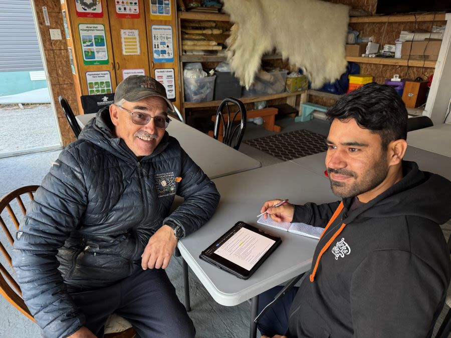Two people sit at a table. One has their elbow on the table and is smiling at the camera. The other is sat closer to the camera and looking past it. They have a tablet and pen and paper in front of them.