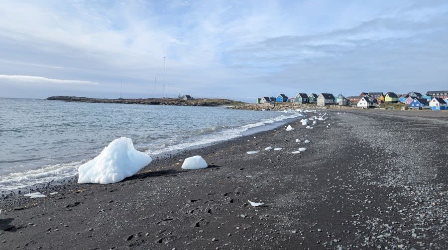 A rocky beach with large chunks of ice littered on the shoreline.Brightly-coloured houses are dotted along the coastline. 