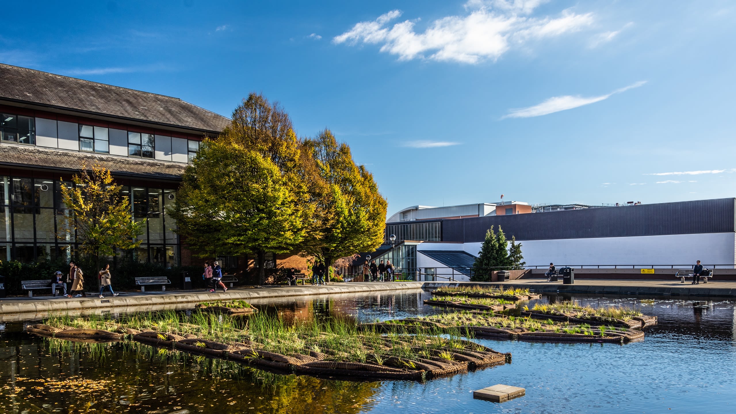 The Roger Stevens Pond, shimmering on a sunny day, with University buildings in the background.