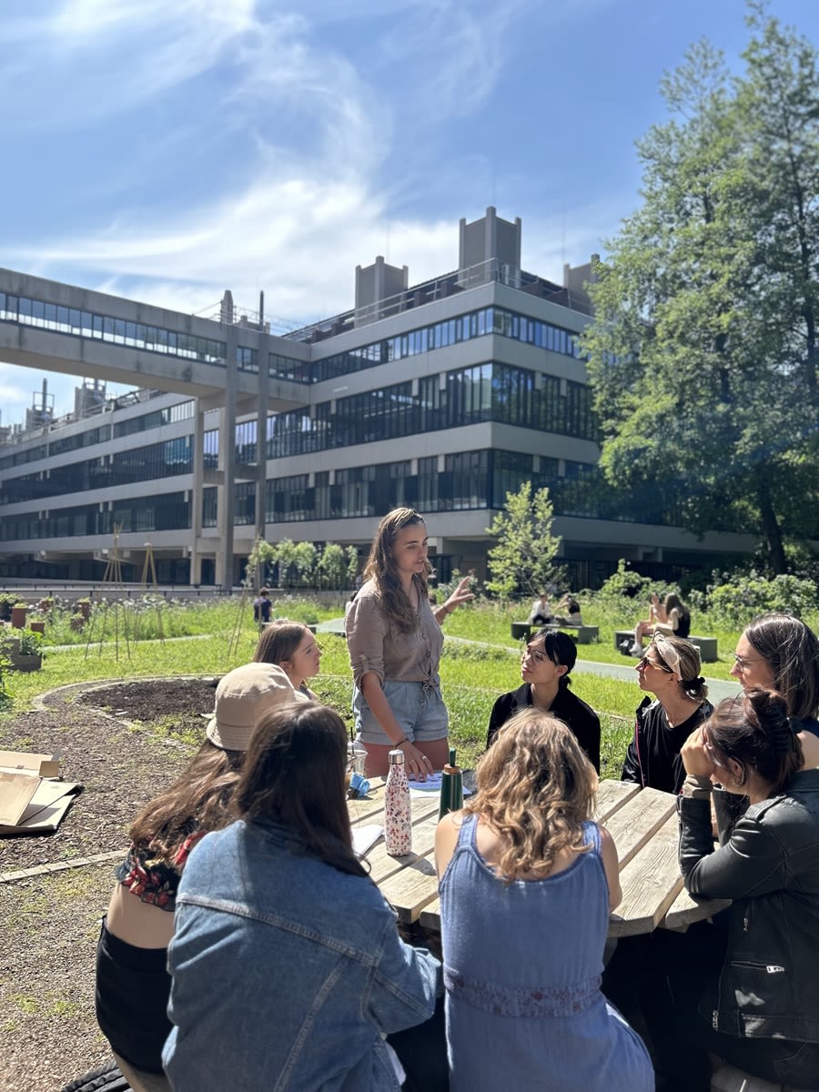 A group of students sat around a park table, listening to another student who is stood up, talking. The sustainable garden is in the background.