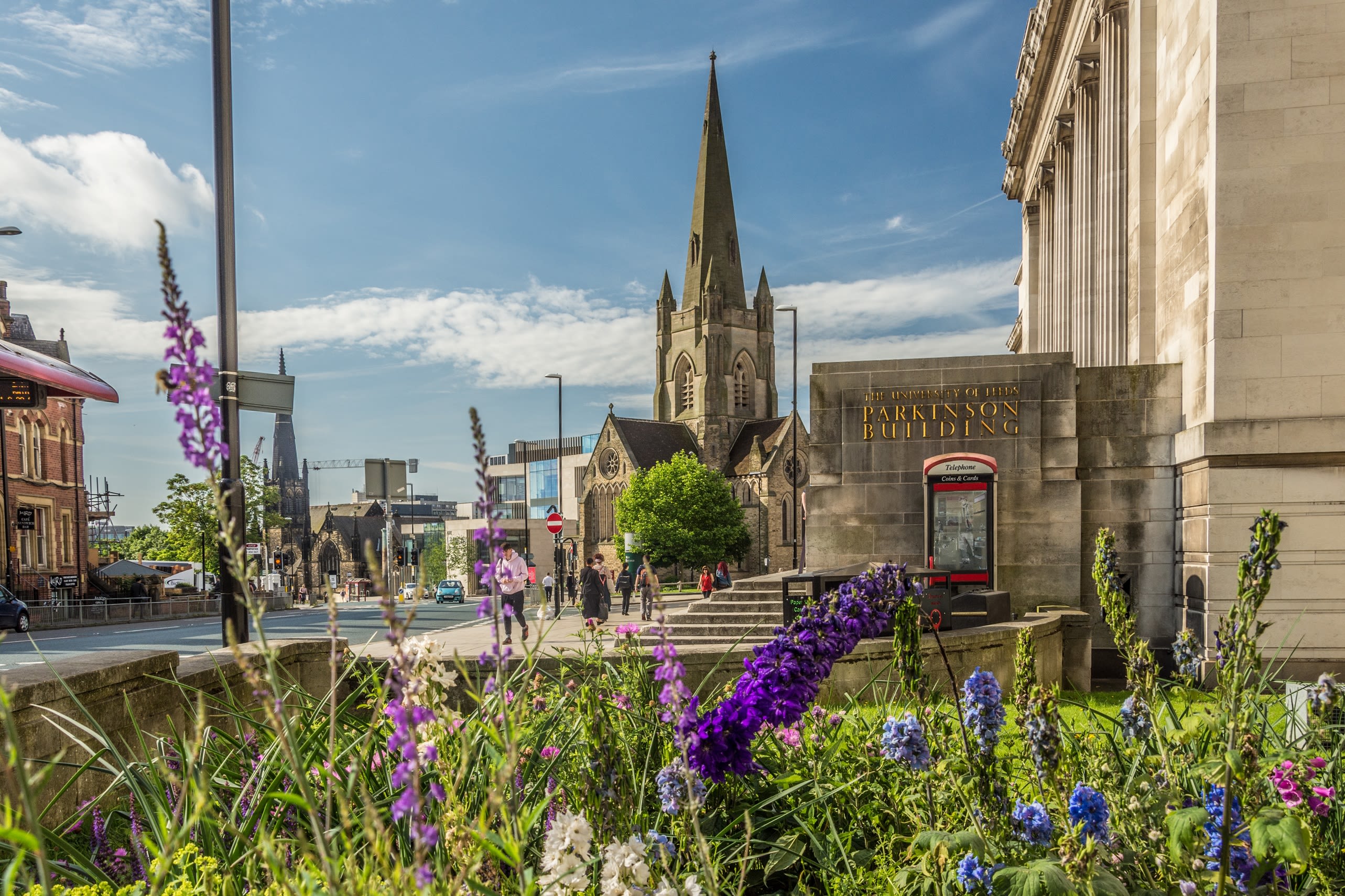 A sunny street with blue sky, showing part of the Parkinson Building on the right, flowers and plants in the foreground and a church spire in the background.