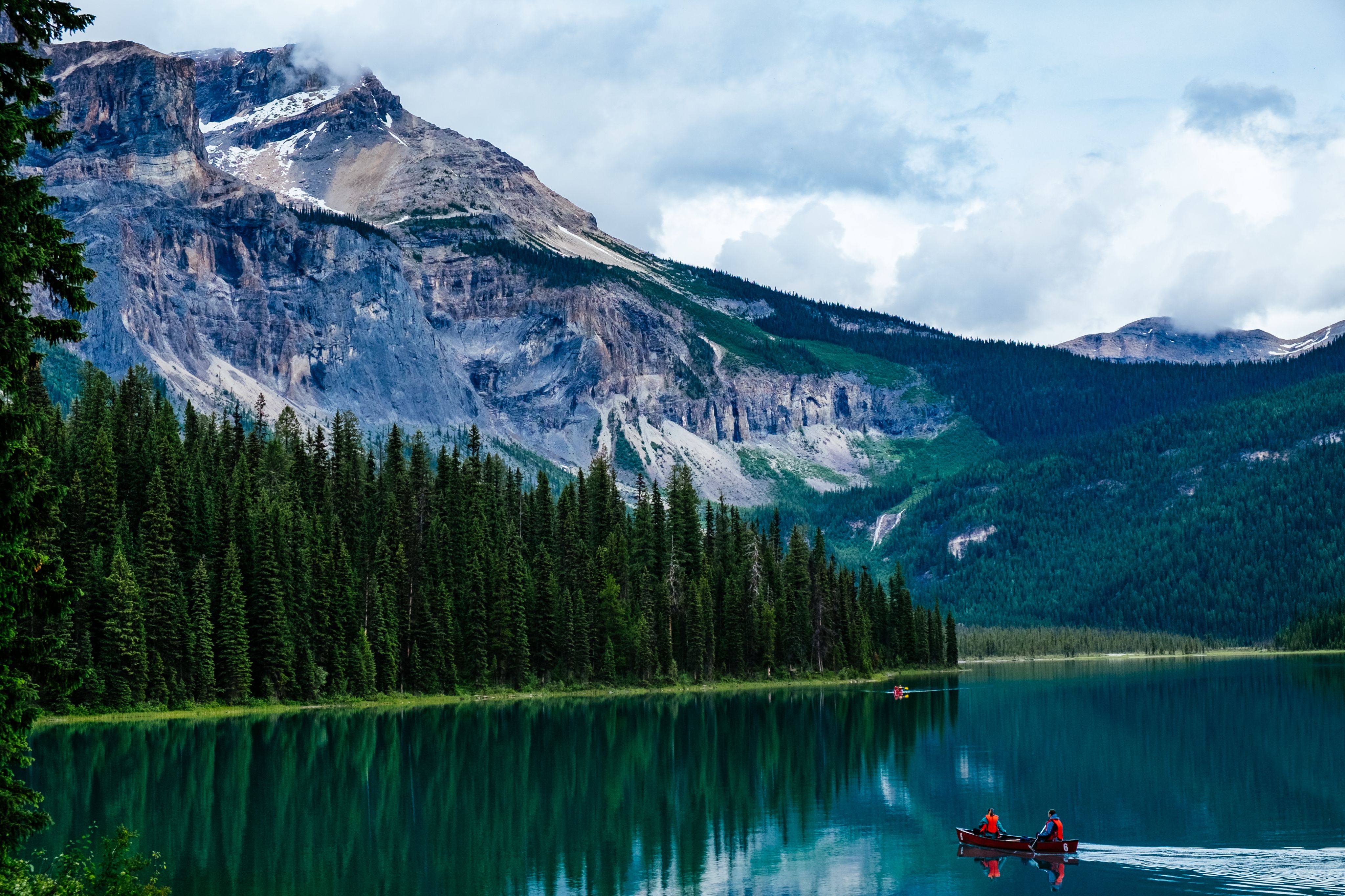 Emerald Lake with a pine forest and the Canadian Rockies behind. A canoe with two people in bright orange life jackets glides on the water.
