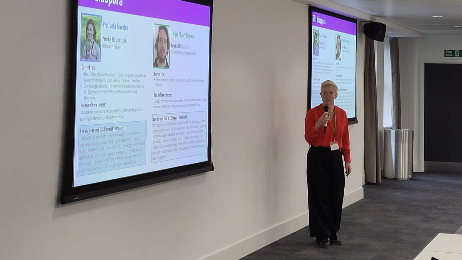 Alice Owen, Director of SRI, stands in front of a presentation, speaking to a microphone. The presentation has pictures of SRI alum.