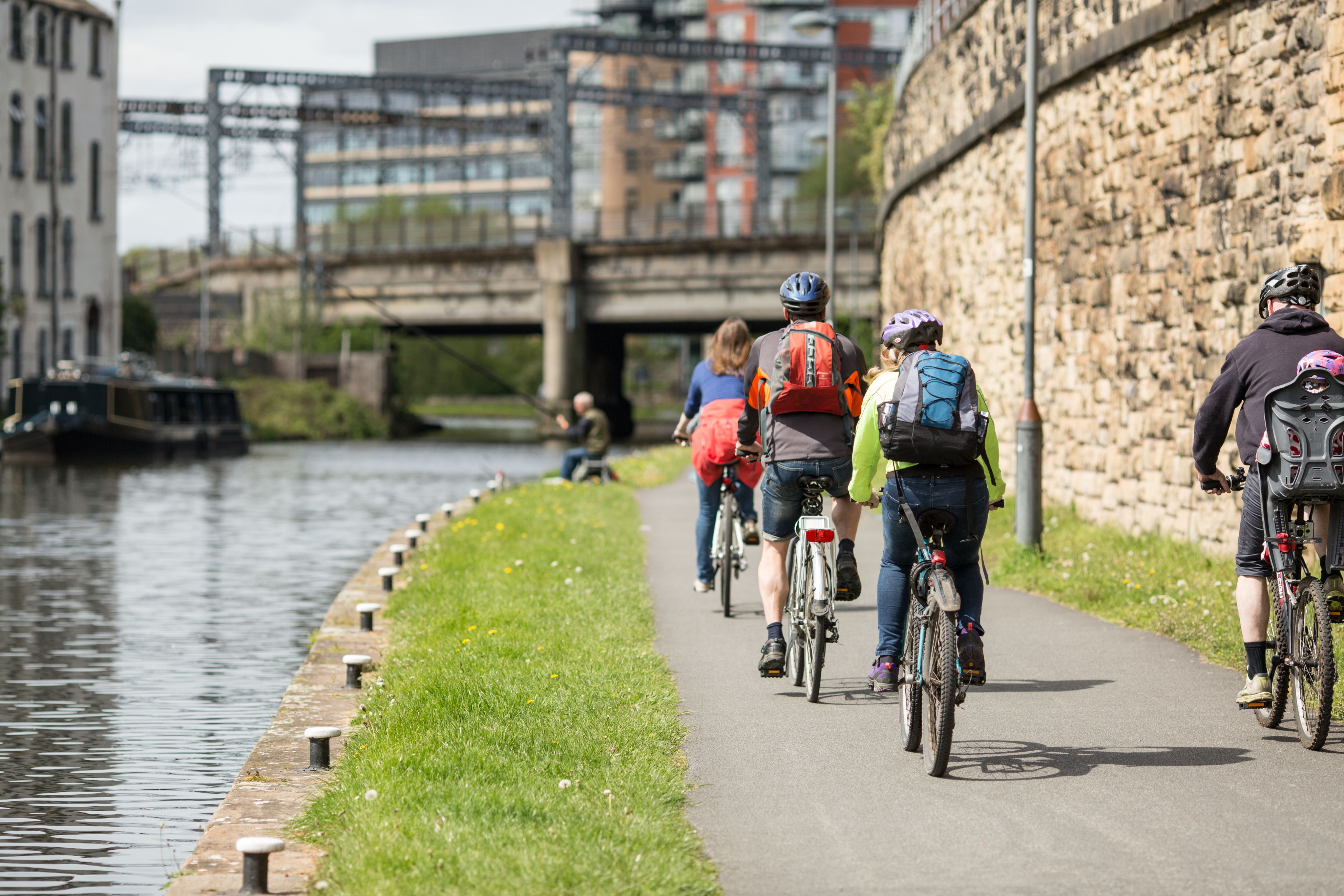 Three cyclists on the path next to the Leeds-Liverpool canal. In the background are a canal boat and a person fishing.