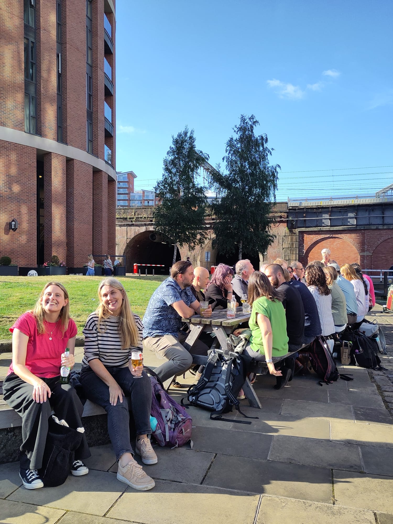SRI researchers sitting on two picnic benches, chatting with drinks on a sunny day. Two people face the camera, smiling.