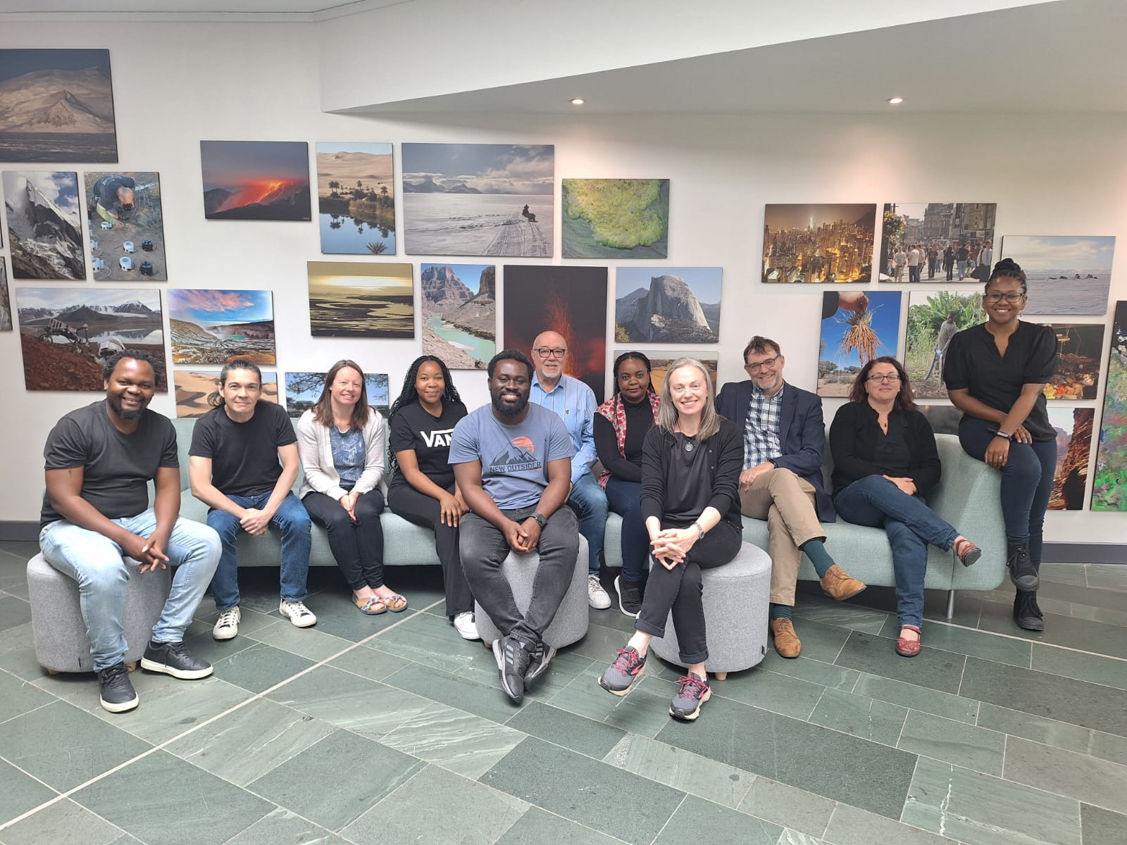 A group of academics from Leeds, York and Pretoria, sitting on stools and smiling towards the camera. The wall behind is filled with pictures of landscapes from around the world.