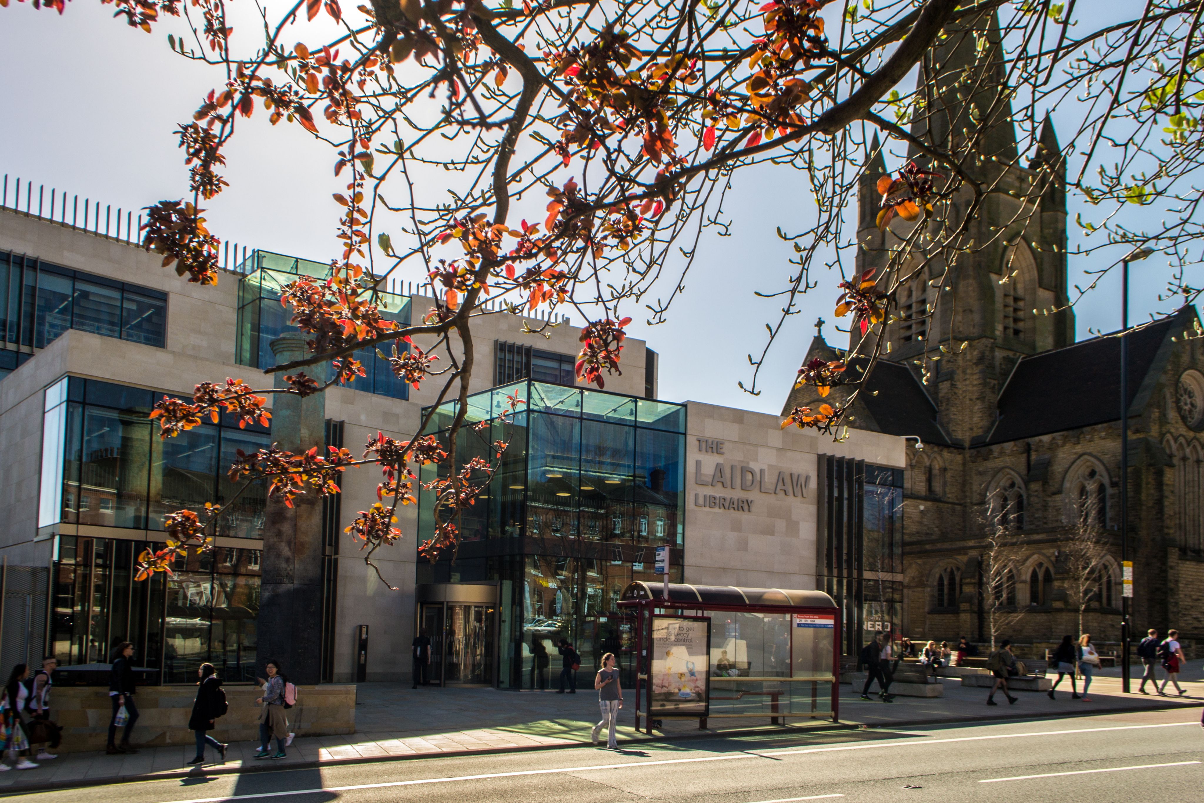 The Laidlaw Library. It is a grey stone building with lots of glass windows. There is a pavement, some trees, a bus stop, and some people walking around outside.