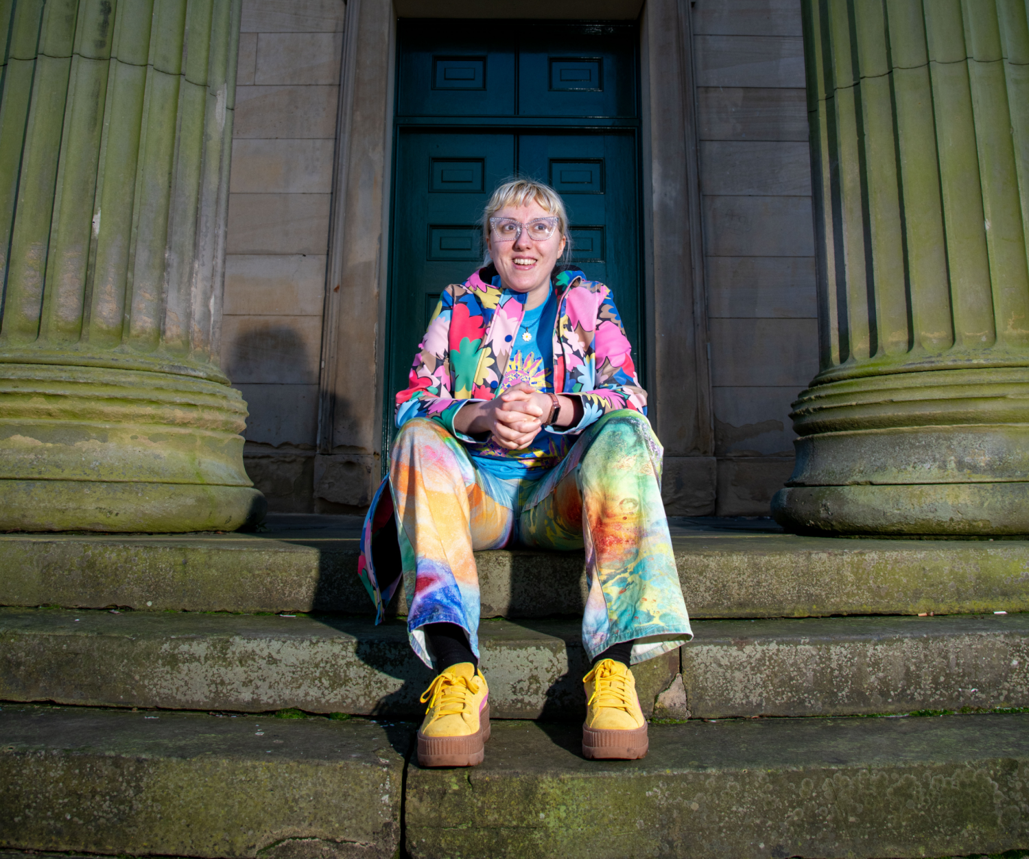 Kay Child sits on steps before a large brown door, smjling