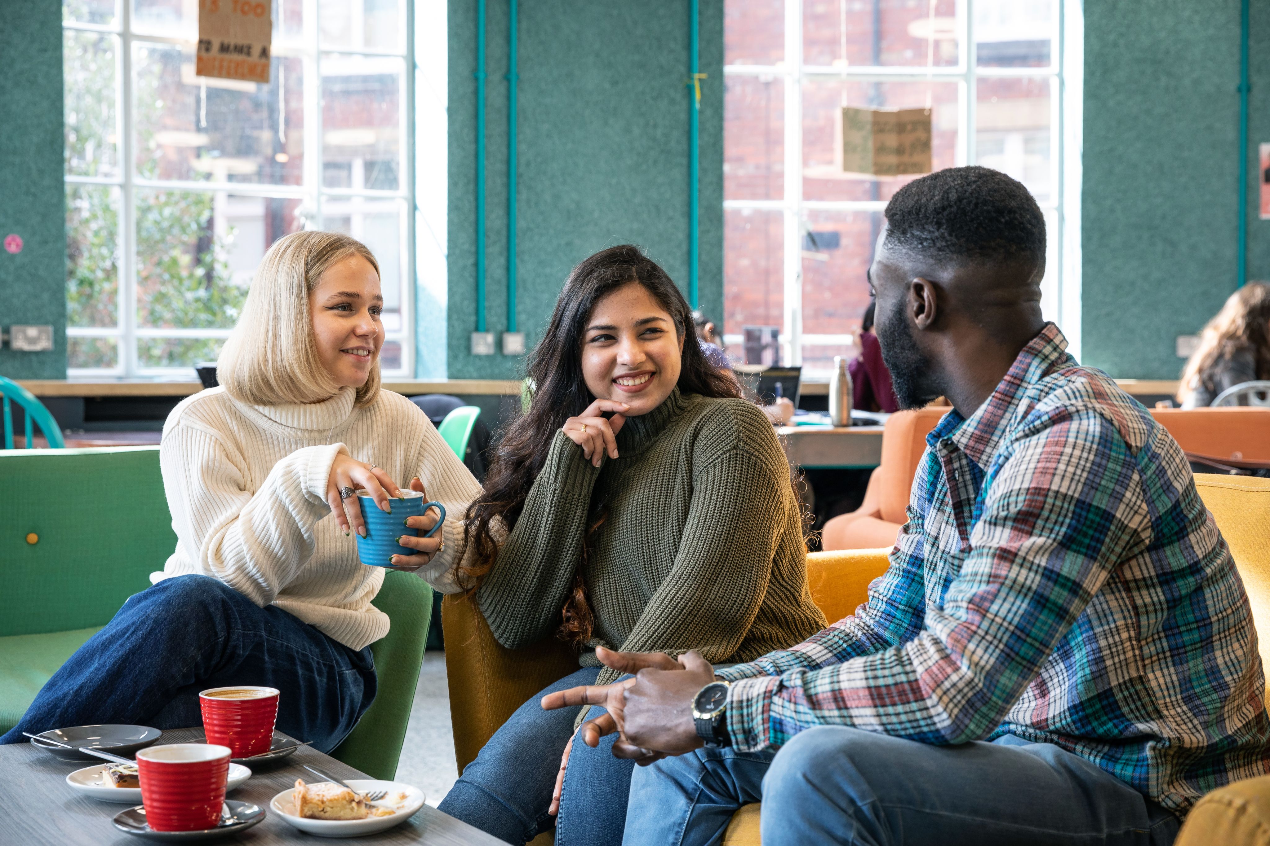 Three students are smiling and talking to each other on green and yellow seats. They have hot drinks and food in front of them.