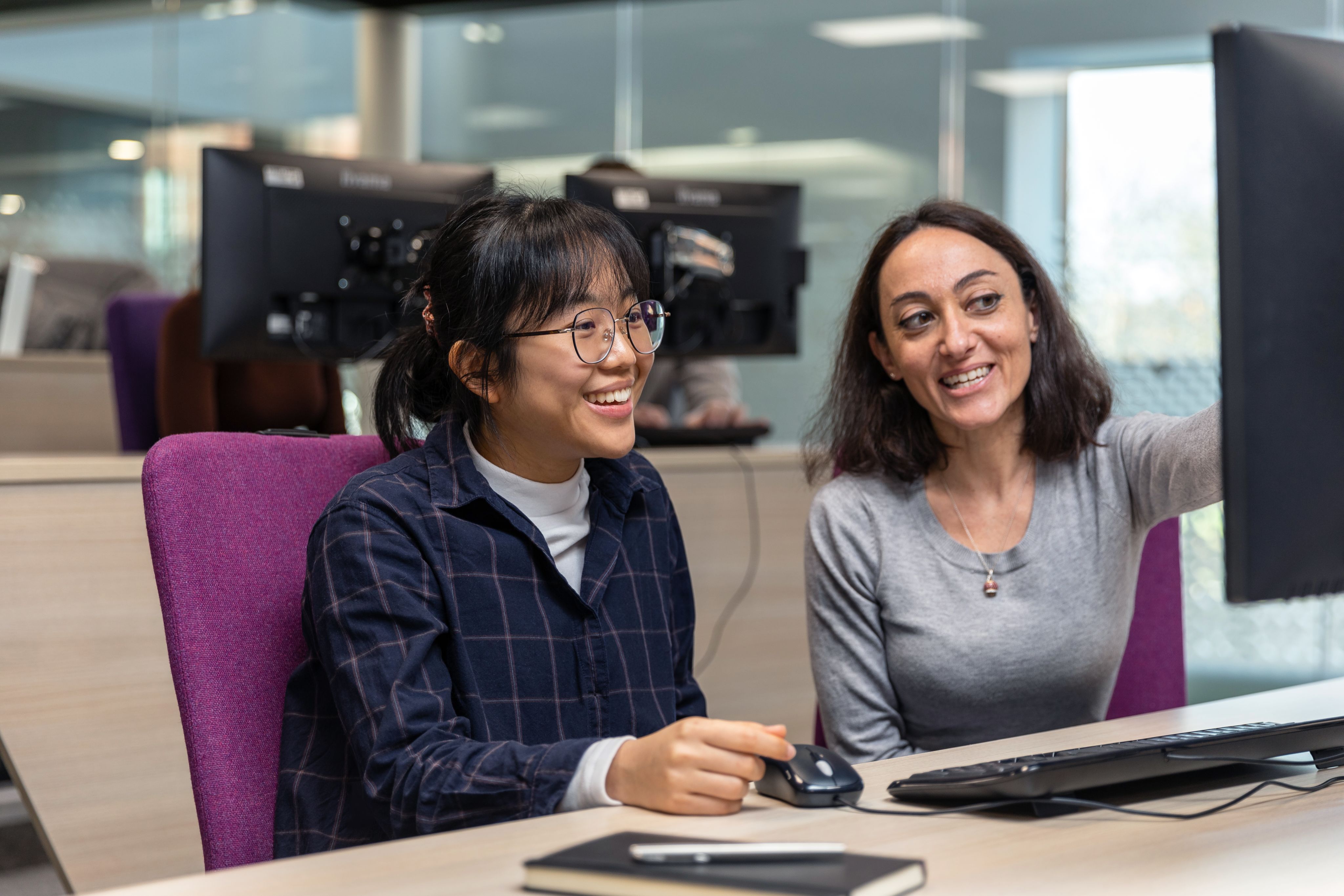 A student and an authority figure are looking at a computer screen together. The authority figure is pointing at the screen. The student has her hand resting on the desk and is smiling. 