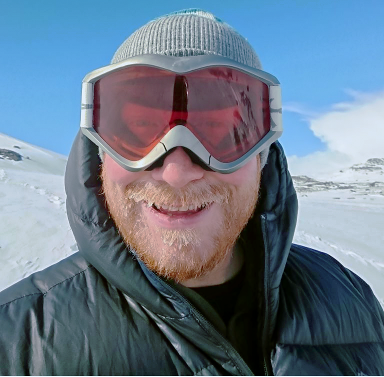male researcher in hat and goggles smiling at the camera while on fieldwork in the arctic.