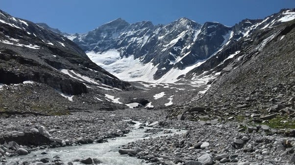 small stream winds through a rocky river bed, with snow covered ravines and mountain tops in the distance.