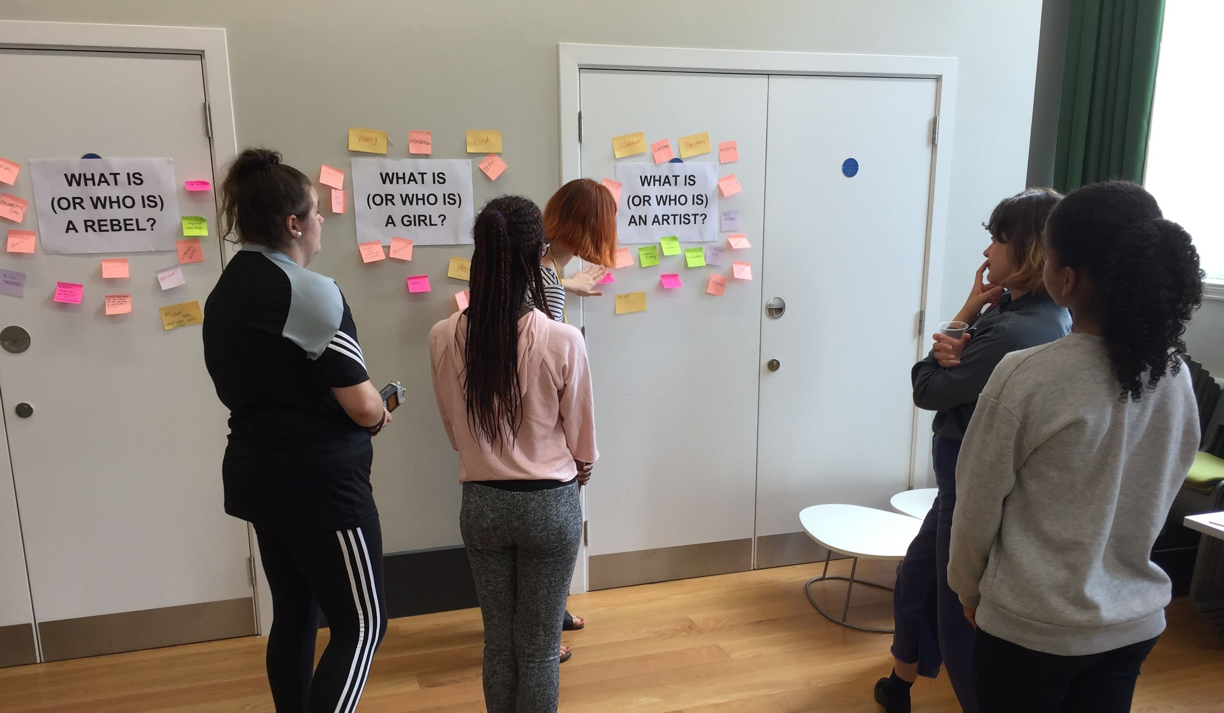 5 people stood in a room looking at sticky notes surrounding three statements. The statements read 'What is (or who is) a rebel?', 'What is (or who is) a girl?' and 'What is (or who is) an artist'?.