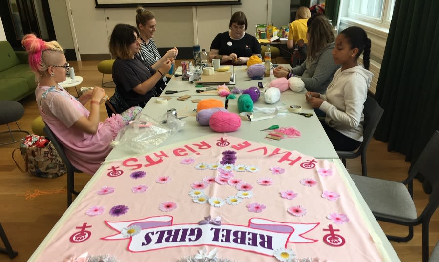 6 people sat at a table making a banner at the Art School for Rebel Girls. The pink banner reads 'Rebel Girls have rights'.
