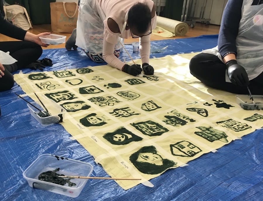 Three people sat on a mat painting faces and buildings in squares on yellow fabric.