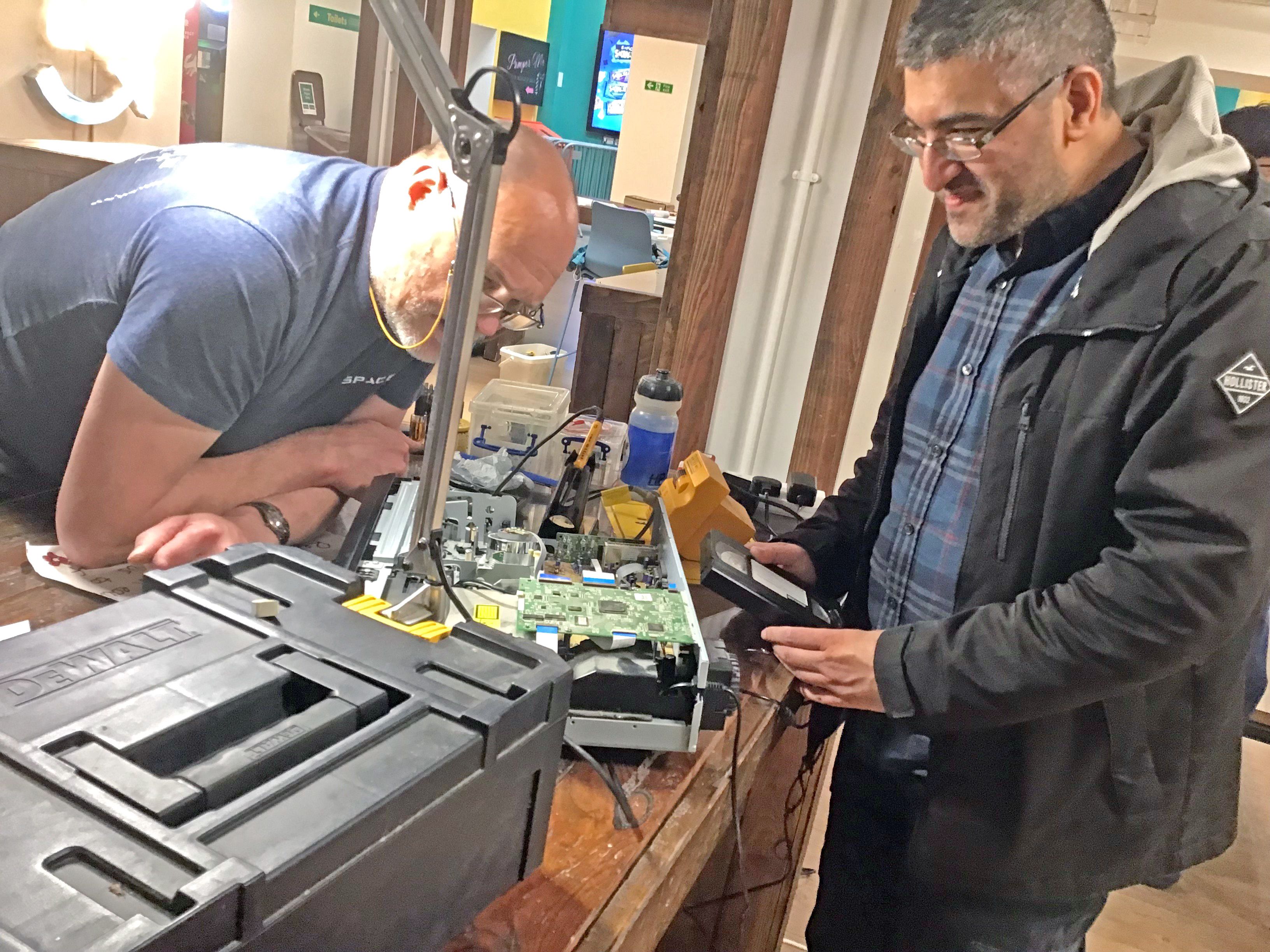 Two people have removed the cover of a large VHS player and are fixing some circuits inside.