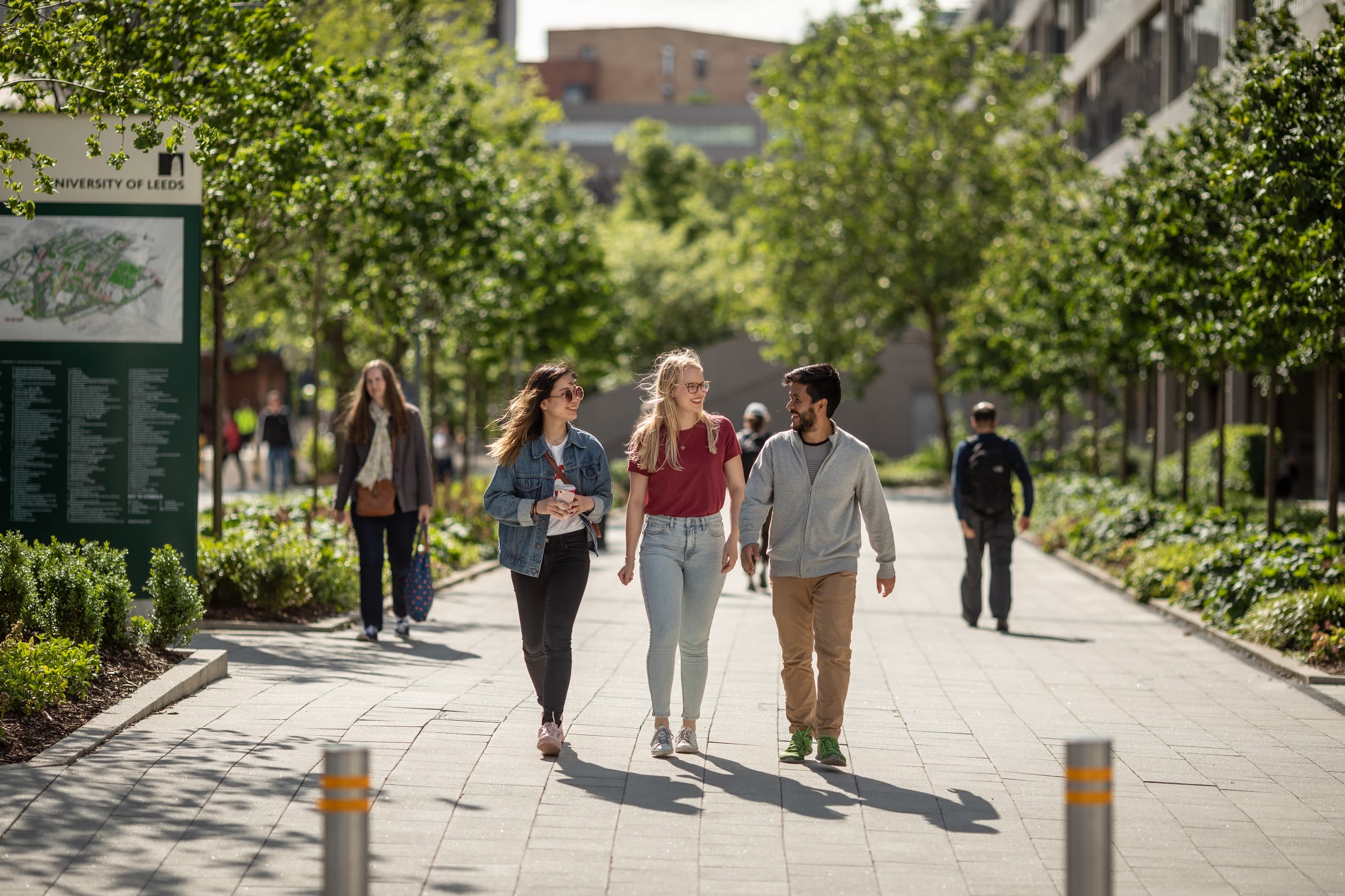 Three students in conversation walking together on a path on the University of Leeds campus. Two people are walking in the background.