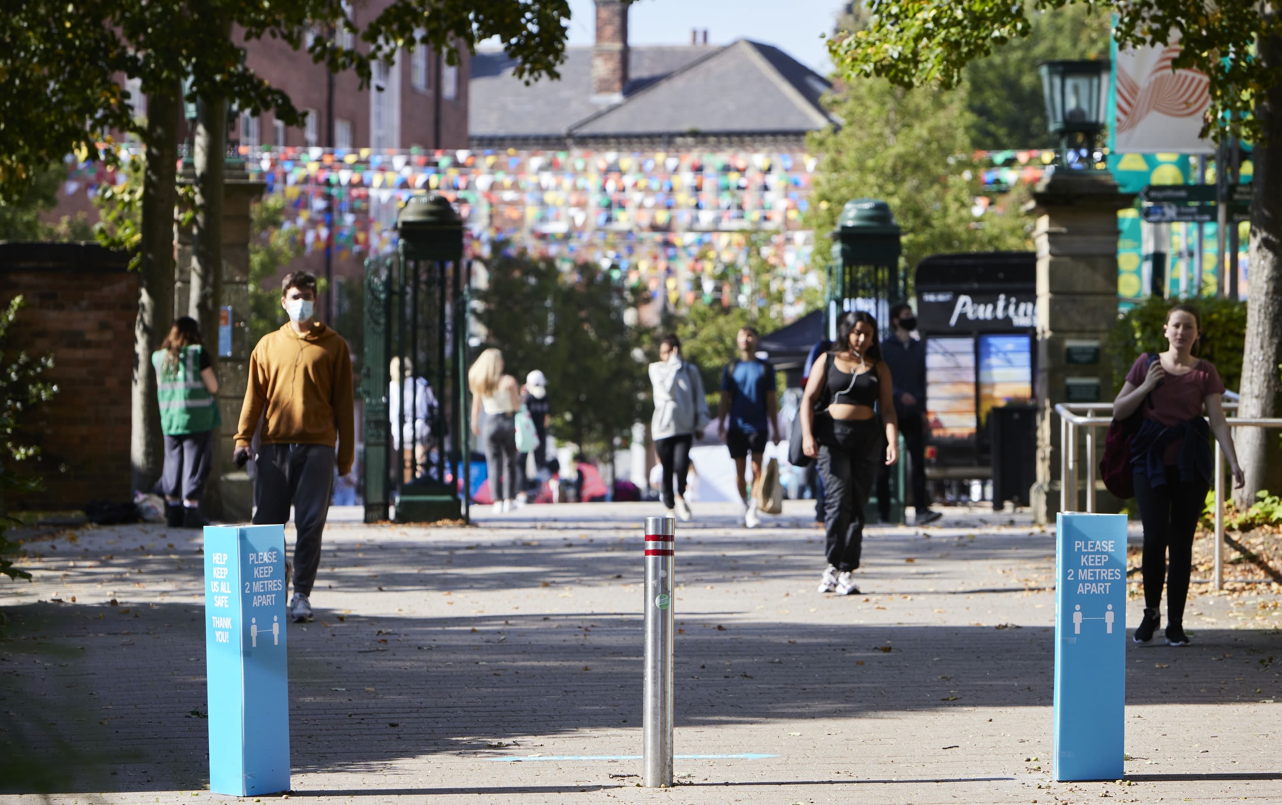 Three people walking outside Leeds University Union, one wears a face mask. There are signs that say 'Please keep 2 metres apart'.
