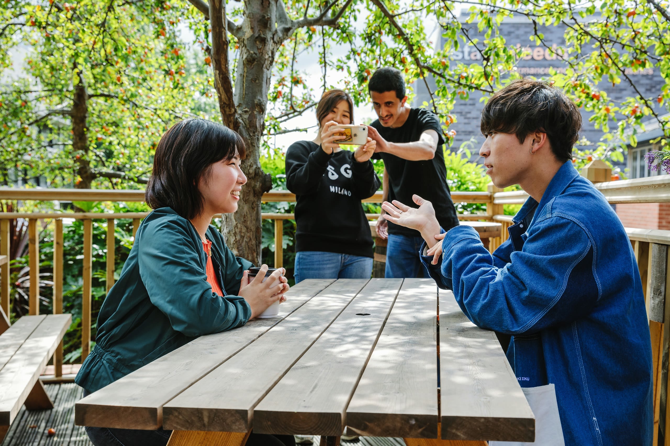 Two people are sat opposite each other on a picnic bench talking. Two more people are talking a photo of them using a mobile phone camera.