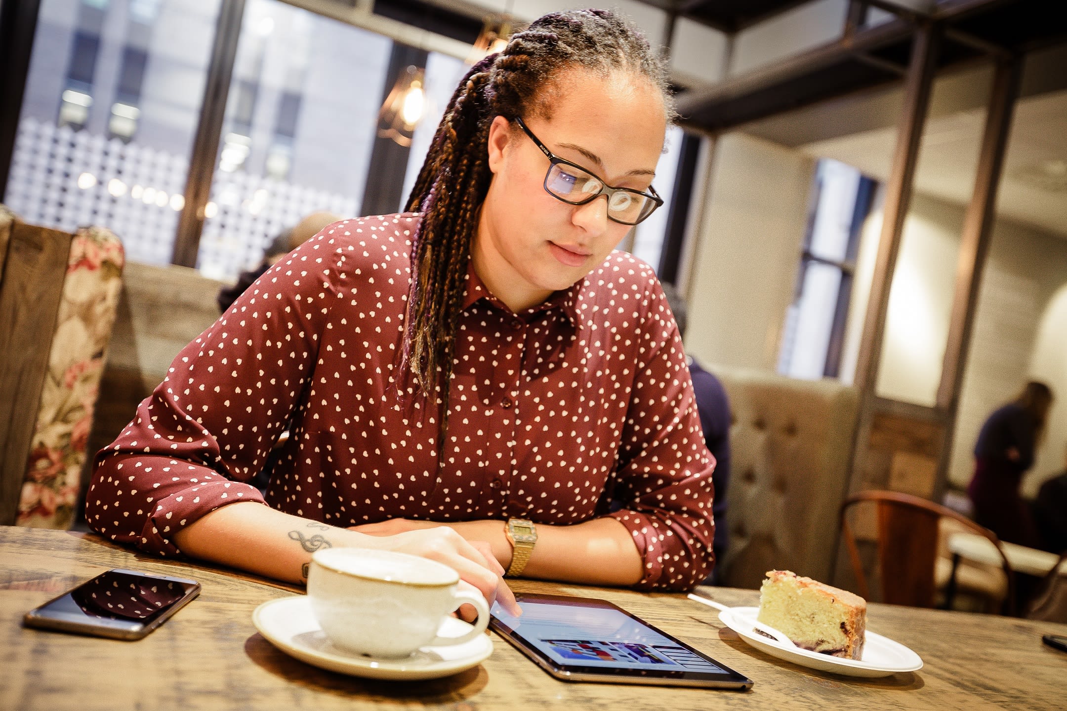 A person wearing dark glasses and a brown polka dot shirt is sat at a table using a tablet. There is a mobile phone, a coffee cup and a slice of cake on the table.