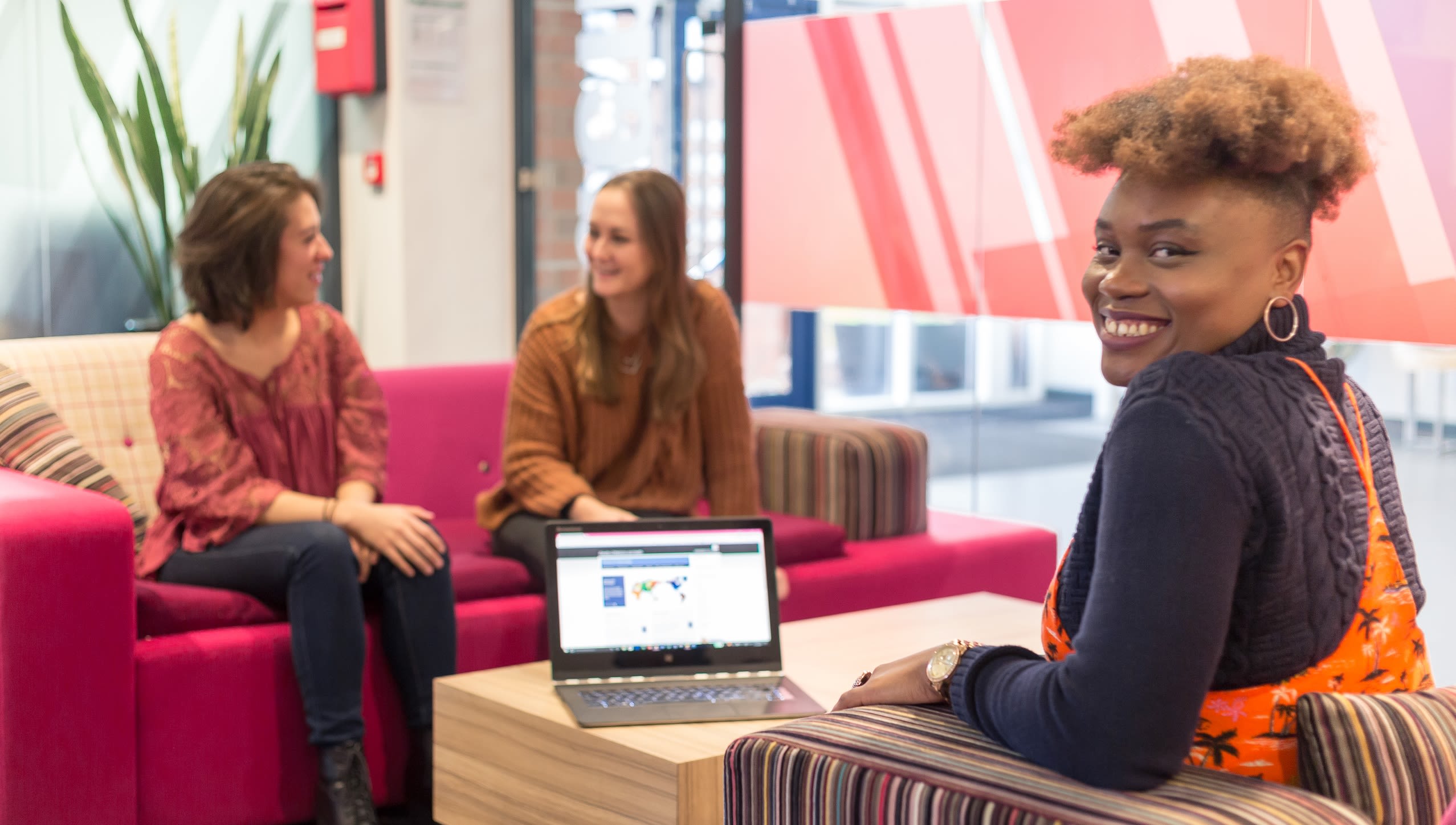 Three people sit on sofas, two are talking, one is smiling facing the camera. There is a laptop on the table in front of them.