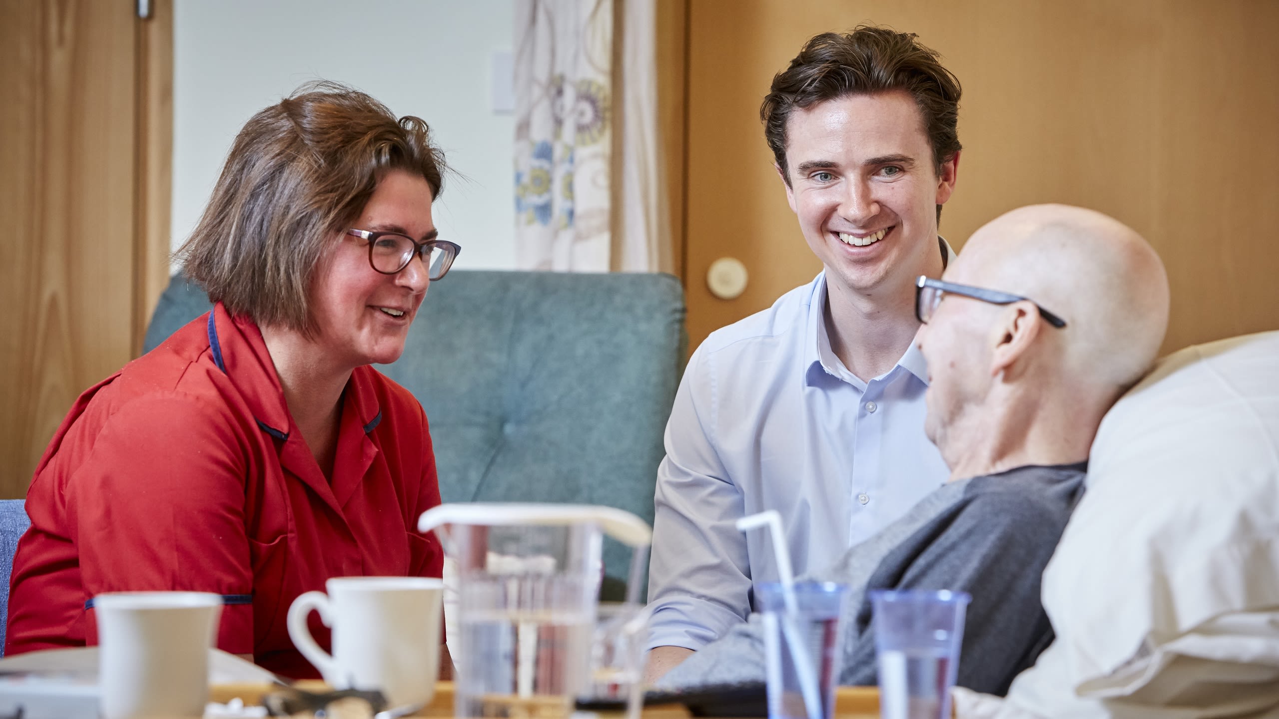 Two members of staff chatting to a smiling patient sat up in a bed in a care setting.