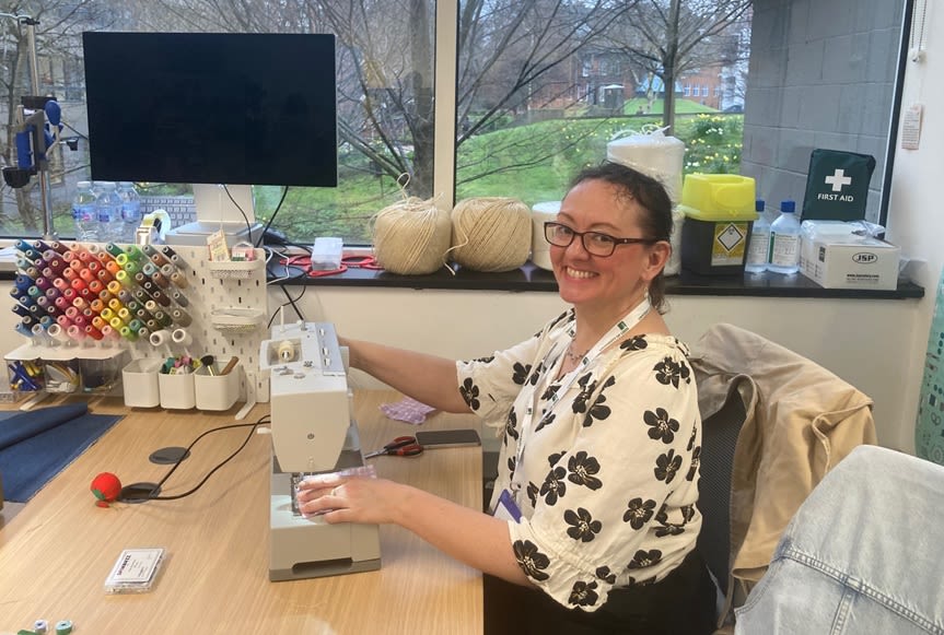 A smiling person using a sewing machine at a desk, with coloured yarns on a nearby shelf. 