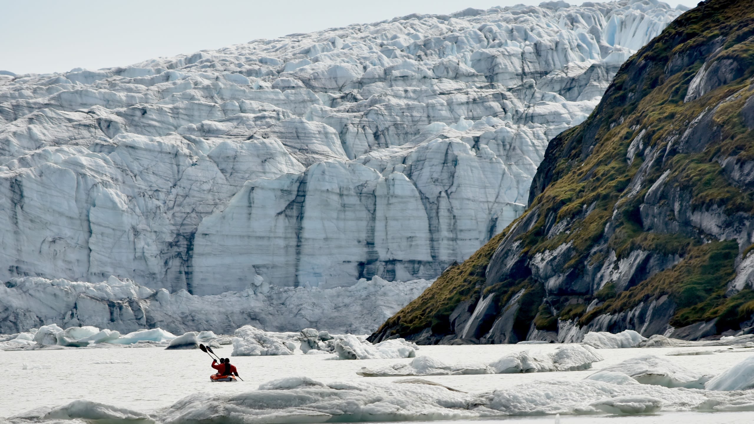 A glacial landscape with huge cliffs of rock and ice and two people on water in a canoe.