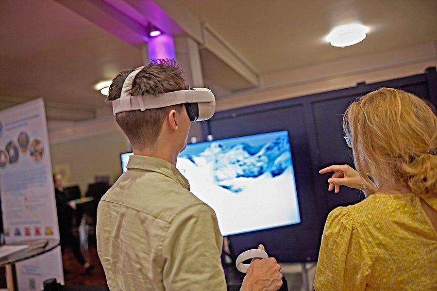 A person talking to another person who is wearing a virtual reality headset. They are facing a plasma screen showing an icy mountainscape.