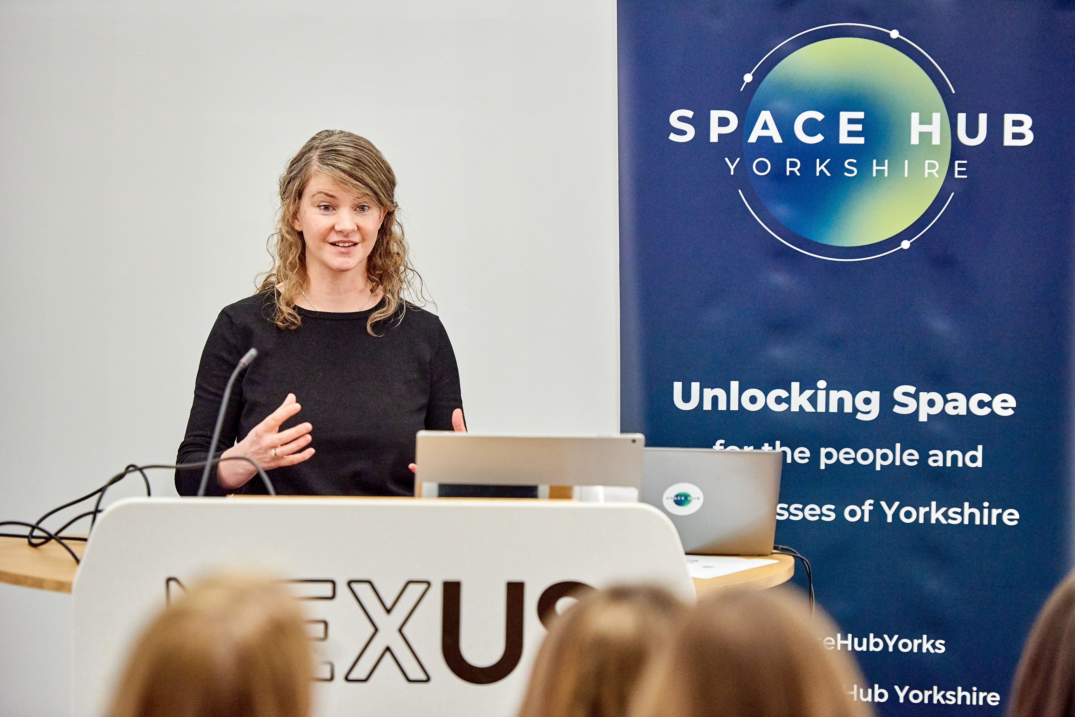 Professor Anna Hogg speaking at a podium. A banner to the right says 'Space Hub Yorkshire' on it