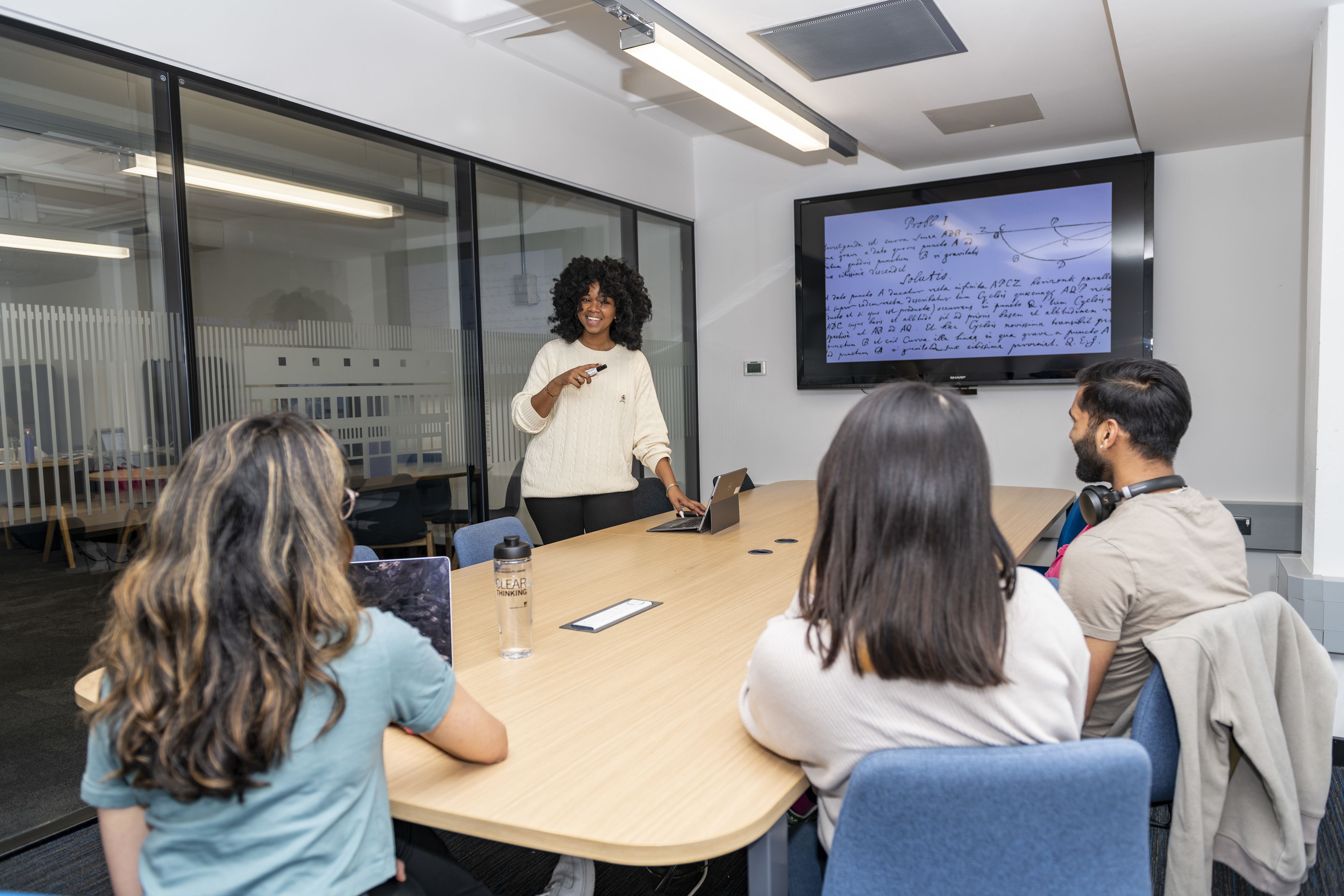 A group sit around a table looking at a presenter next to a large screen